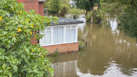 A home surronded by flood water. The water is brown and murky. Trees can be seen around the property.