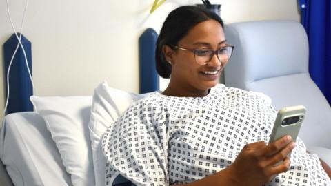 A woman wearing glasses laying on a hospital bed in a gown. She is holding a smart phone in her right hand and reacting happily to what she is looking at one the screen in front of her.