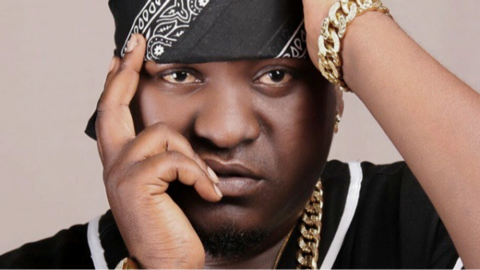 Close-up of musician Sefu Shabani in a black bandana with a white design posing with his hands near his face. He is wearing a chunky gold chain necklace and bracelet.