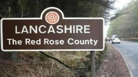 Brown road sign which reads 'Lancashire The Red Rose County' in white letters on the side of a road