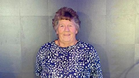 Jo is stood infront of a grey fabric sheet of fabric, looking directly at the camera with a bright smile on her face. She is wearing a blue and white floral print top, and has brown hair with grey streaks towards the sides of her head.