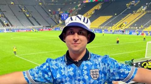 Karim Goodchild with his arms spread out, wearing a blue England shirt and hat, standing in a stadium with a football pitch behind him