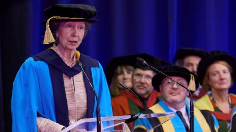 The Princess Royal is stood up at a lectern wearing a long blue cape and a black hat with a hanging yellow tassel. In the background are several people sat down looking towards her. They are all wearing similar black caps.