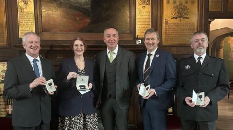Five people dressed smartly and looking directly at the camera. They are smiling and four of them are holding medals. 