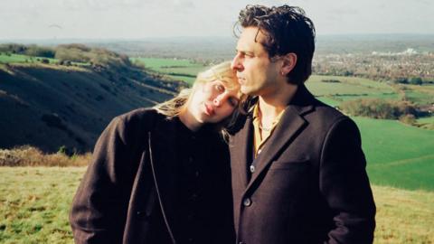 An image of a blonde woman who is wearing all black leaning on a man with brown hair who is also wearing all black in the middle of a field.