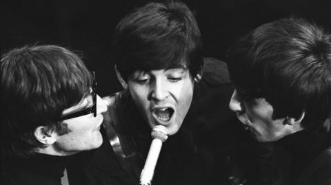 Three Beatles with their distinctive moptop haircuts in monchrome from left: John Lennon with thick black spectacles, Sir Paul McCartney singing directly into the microphone (centre) and George Harrison singing in a side profile.