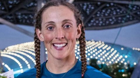 A close-up image of a woman with brown hair in two plaits, wearing a blue t shirt and smiling at the camera