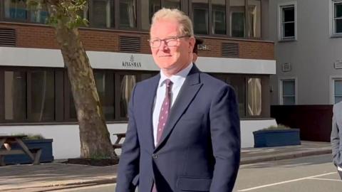 Andrew McLaughlin, the chief executive of the Government of Jersey, walks along a street in St Helier on a sunny day while wearing a navy blue suit, white shirt and maroon tie. He is wearing glasses and has trimmed blond hair. An office building for a firm called Kingsman is on the other side of the road along with a tree, two benches and two planters.