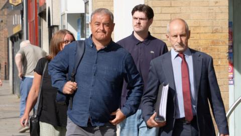 David Moore and other members of Emily's family walk, along with a bald bearded man in a suit carrying a large ring binder of documents, into the coroners court building in Crook. David Moore is clean shaven with short grey hair and a determined expression on his face. He is wearing a dark navy shirt. Behind him, partially seen, is a woman with shoulder length dark brown hair wearing glasses and a black short-sleeved top. Behind the suited man, who is on David's left, is a younger man with short dark hair in a centre parting. He has a moustache and wears a dark - perhaps purple - polo shirt and stonewashed blue jeans.