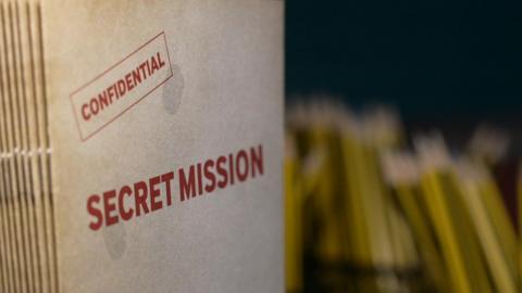 A manila folder on which 'Confidential' and 'Secret Mission' are written. There are other folders stacked up behind this folder.