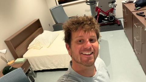 A man with a criffy beard wears a grey t-shirt and smiles as he takes a selfie in a room with a bed and exercise bike in the background