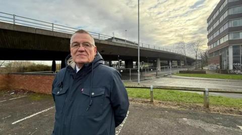 Martin Gannon standing in a car park near the Gateshead flyover. He has short hair and is wearing glasses and a navy coat. The concrete bridge is behind him. Temporary fencing has been put up across the road running under the bridge.
