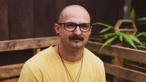 A bald man with glasses, earrings and a moustache wearing a yellow shirt