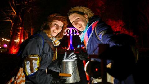 Two people in colourful costumes are smiling at the camera. A light installation is illuminated behind them.