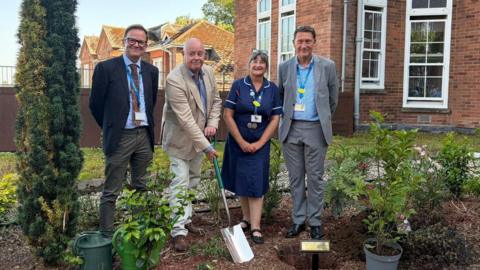 Four people in a garden planting a tree with a brick building behind them.