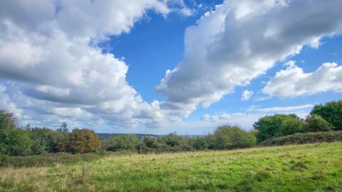 A sunny day looking out over a field lined with trees. The sky is blue but filled with some white clouds. Hills can be seen in the distance