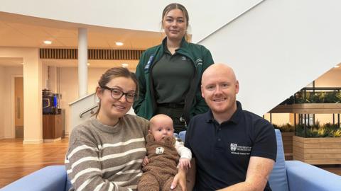 A woman wearing a green paramedic's outfit stands behind a blue sofa in a large room. Sitting on the sofa are a man, a woman and a small baby in the middle, who they are both holding.