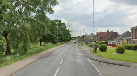 A straight section of Crosshall Road with houses on the right and trees and grass on the left of the road. 