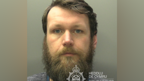 Carl Martin looks straight ahead at the camera, wearing a grey jumper and blue top. He has blue eyes and a beard. The South Wales Police logo can be seen in the photo.