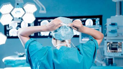 A healthcare worker in blue surgical scrubs stands in an operating theatre, tying the straps of a disposable surgical cap. Bright operating lights hang overhead, while illuminated medical scans and imaging screens are displayed on monitors across the room. An operating table covered with blue drapes and additional medical equipment can be seen in the background.
