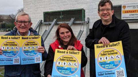 Two men in dark coats are standing either side of a shorter woman in a red gilet. They are all holding blue and yellow signs promoting the scheme.