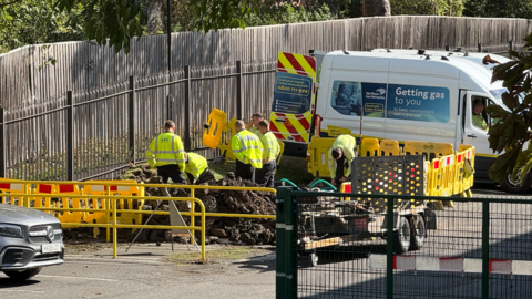 Gas workers, dressed with high visibility jackets, are standing next to a mound of soil from underneath the school's car park. In the distance is a white van which says 'getting gas to you', and in the front, there are several yellow barriers.