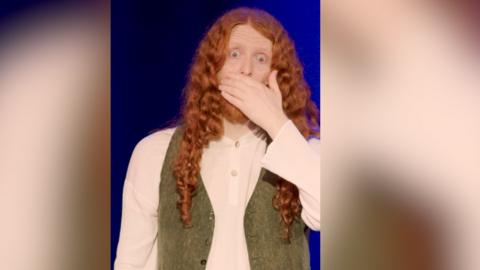 A man with long red hair and a green waistcoat clasps mouth over hands in shock.