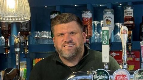 A man wearing a green top is standing behind a pub bar. He has short fair sweptback hair and a grey-tinged beard.