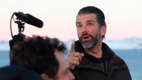 Donald Trump Jr, wearing a green jacket, points up while he is speaking. There is a microphone device in front of him, suggesting he is being filmed as he speaks.