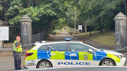 A police car and police tape outside a street lined with trees.