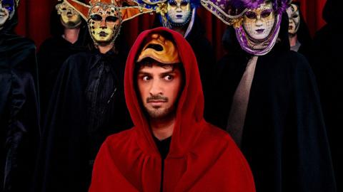 Shabaz dressed in a red cloak. Behind him is a group of people dress in black cloaks wearing masquerade masks