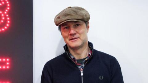 David Morrissey smiles at the camera in 鶹Լ Studios, wearing a brown flat cap and Fred Perry navy jacket