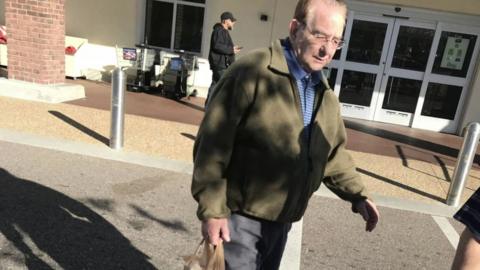 A man with thinning hair, glasses, a blue shirt, and a green fleece walks across a car park. He looks down at the ground