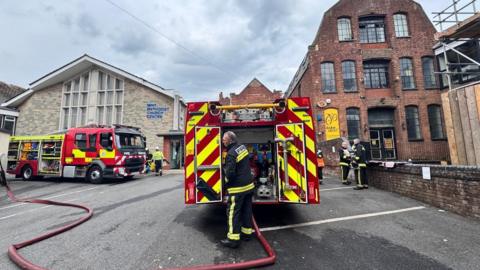 Fire engines and firefighters at the scene of a substation fire in Exeter. There are hose reels on the ground and fire crews dotted around in the background.