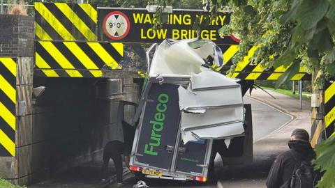 A van is partially destroyed after hitting a railway bridge. The roof is shredded and a man can be seen looking at one of the rear tyres. Another man is seen on the pavement looking at the scene.