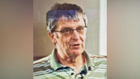 A head and shoulders image of a man wearing a striped polo shirt and glasses. He looks as if he is talking