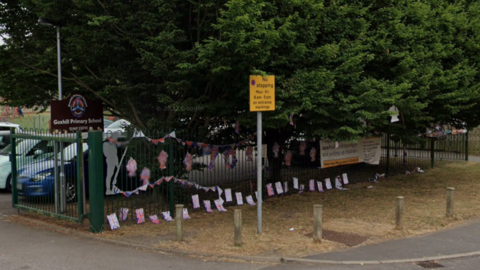 The photo has been taken from the street outside a primary school. There is a green fence just off the footpath, which has union jack bunting hung on it. To the left is a brown sign which reads 'Goxhill Primary School.' The are multiple cars parked in a car park behind it.