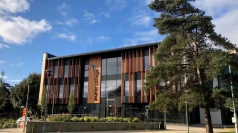 A picture of the front of Exeter College. There is a number of trees and hedges in front of the building with is made out of glass and wood. 