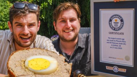 Two men, both with brown hair and short beards, are seated at an outdoor table holding a Guinness World Records certificate and are smiling at the camera. In front of them lies a massive Scotch egg, sliced in half to reveal the egg inside.