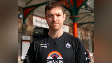 Alex Rigby has short brown hair and is wearing a black t-shirt with Merseyrail written in white lettering and a Centre 56 rainbow-with-clouds logo.