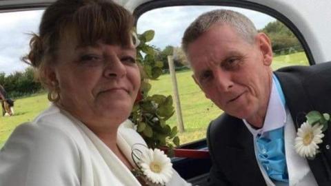 Carol and her husband sitting in a white wedding car, they appear to be in their 50s or 60s. Carol wears a white suit jacket and pink flowery top below, with a white gerbera on her lapel. Her husband had a bright blue kravat, white waistcoat and black suit jacket. In the background is a field with a horse.