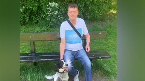 Steven Mead wearing a white T-shirt with a light blue square at the front of it and blue jeans. He is sitting on a park bench and smiling at the camera.
He has a dog sitting next to him.