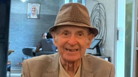 John Mackey smiles for a photo in a cafe. He is wearing a pork-pie hat and a cream jacket and light shirt. 