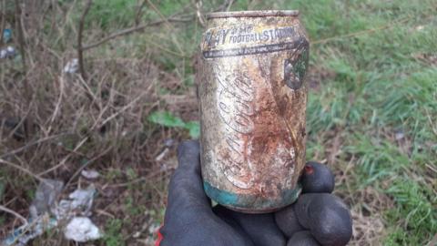 A hand holding a Coke can that is faded. There is more rubbish on the ground in the background.