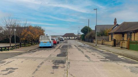 A concrete road, the surface of which is very patchy. There are houses on the road and cars and motorhomes parked up.