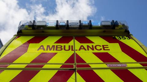 The back of an ambulance taken from the ground looking up. There is diagonal red and yellow stripes, with the word ambulance on the vehicle. Blue lights sit on top. There is a cloudy sky above it.