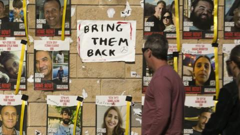 People walk past posters of Israeli hostages held in Gaza and a sign saying "Bring them back", on a wall in Tel Aviv, Israel (20 December 2023)
