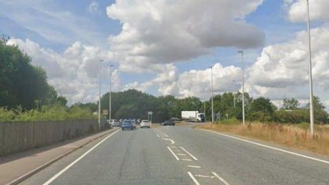 A view of the Wragby Road roundabout near Lincoln. The photograph is taken in the summer and the grass verge is going brown. A fence lines the other side of the road and tall lamp posts are either side of the carriageway.