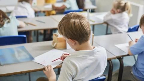 File shot of a school classroom. A boy wearing a white school T-shirt is sitting on a blue plastic chair at a grey desk. He is holding a piece of white paper with his left hand and drawing on it with a red pencil with his right. There is another picture on the table and a wooden box rests in front of him. There is a girl in a blue dress to his right who is also holding a pencil.