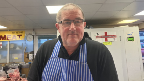 Market trader Gary on his meat stall in Freeman street market, he is wearing a blue and white vertically striped apron. He is standing in front of a fridge and packs of raw meat are on a counter behind him. 